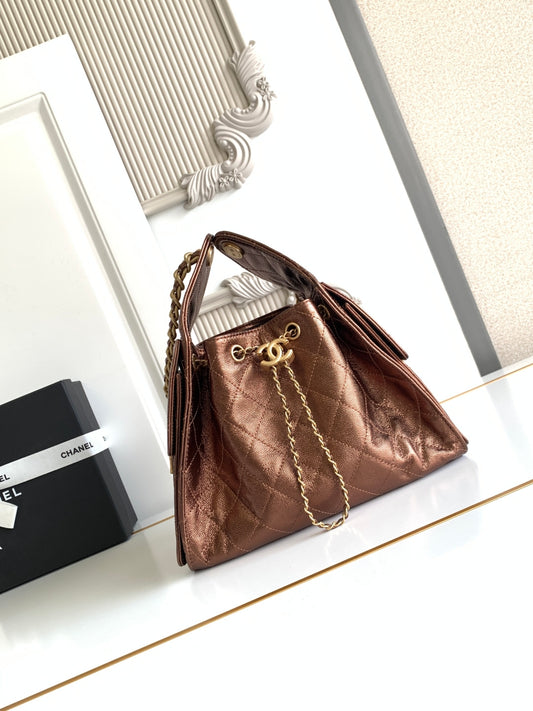 A metallic bronze Chanel bucket bag with a quilted pattern and gold chain strap, displayed next to a black Chanel box on a white surface.