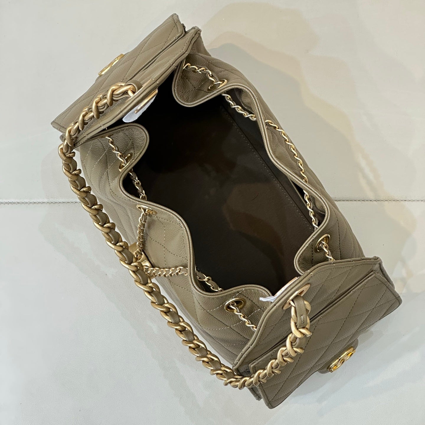 A taupe leather shoulder bag with gold-tone chain strap and hardware, shown from an overhead angle on a light gray surface.