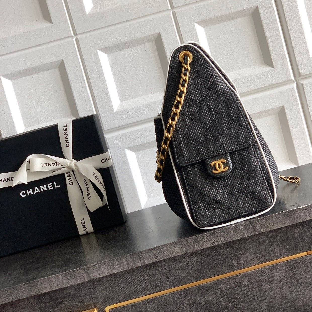 A black Chanel shoulder bag with a textured snakeskin pattern, gold chain strap, and gold interlocking CC logo, displayed next to its black Chanel gift box on a dark gray table against a white tiled wall.