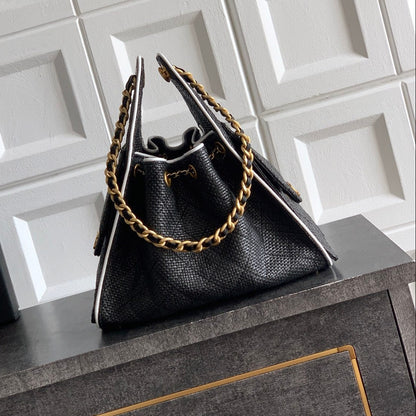A black textured leather handbag with gold chain straps and white piping, displayed on a dark gray cabinet next to a black Chanel gift box against a white tiled wall. Chanel 25 Raffia Black