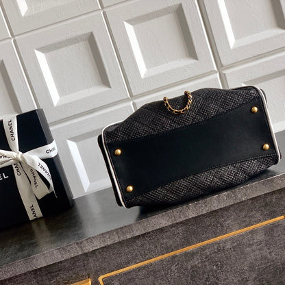 A black Chanel shoulder bag with a textured woven body, a smooth black front panel, gold-tone studs, and a gold chain detail, displayed on a dark cabinet next to its branded black gift box.