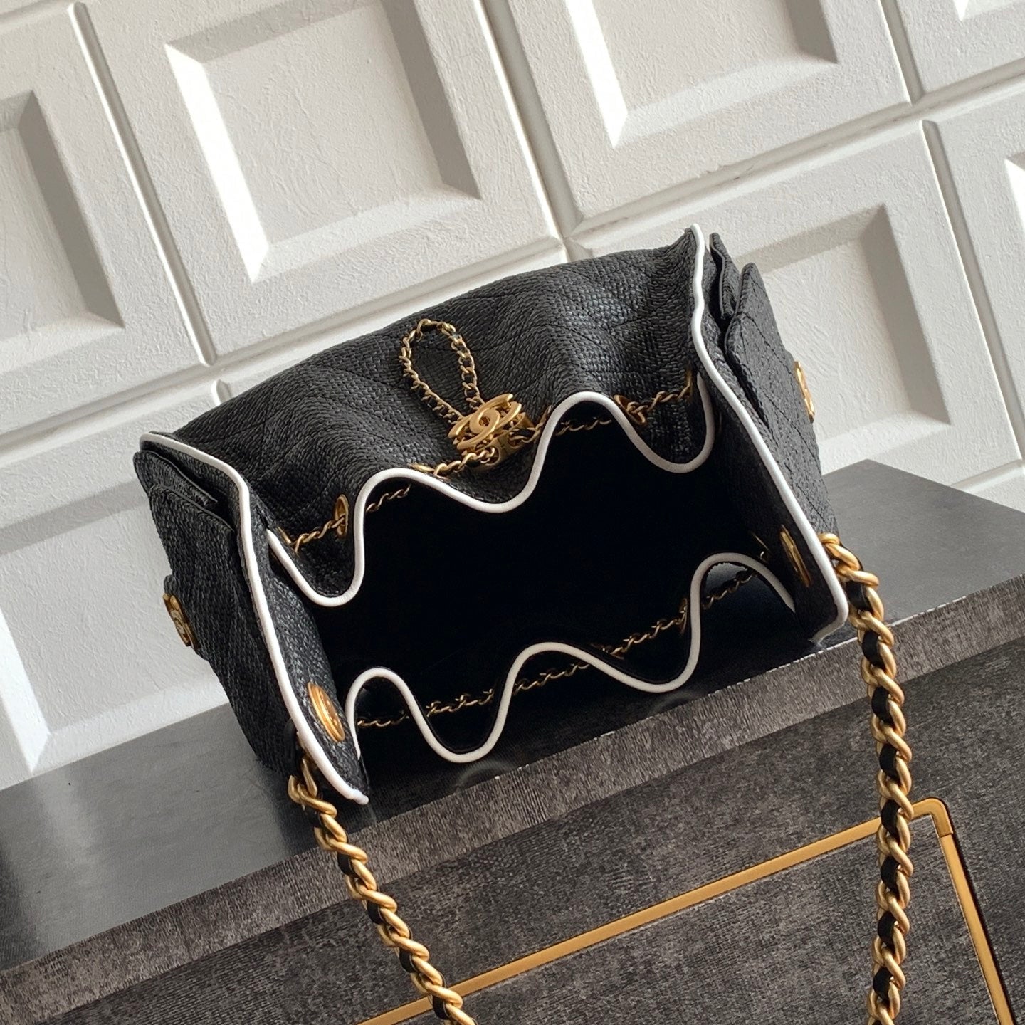 A black textured leather handbag with gold chain detailing and white piping, shown resting on a black wooden surface against a white paneled wall.