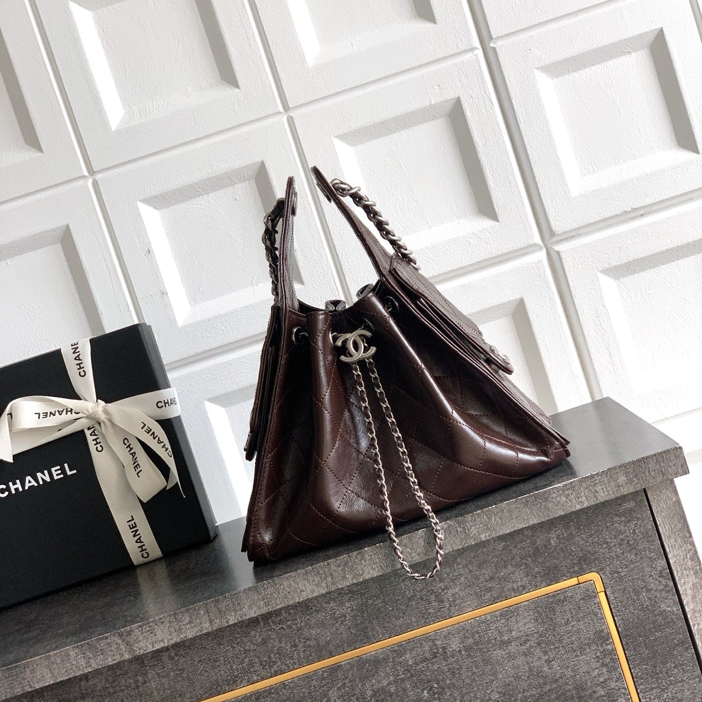 A dark brown quilted leather handbag with silver chain straps and a silver interlocking CC logo, displayed on a dark gray cabinet next to a black Chanel gift box against a white tiled wall.