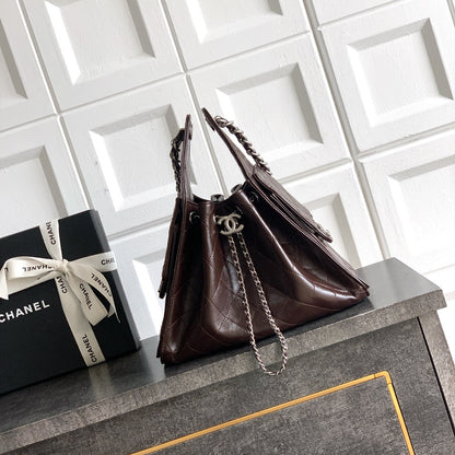 A dark brown quilted leather handbag with silver chain straps and a silver interlocking CC logo, displayed on a dark gray cabinet next to a black Chanel gift box against a white tiled wall.