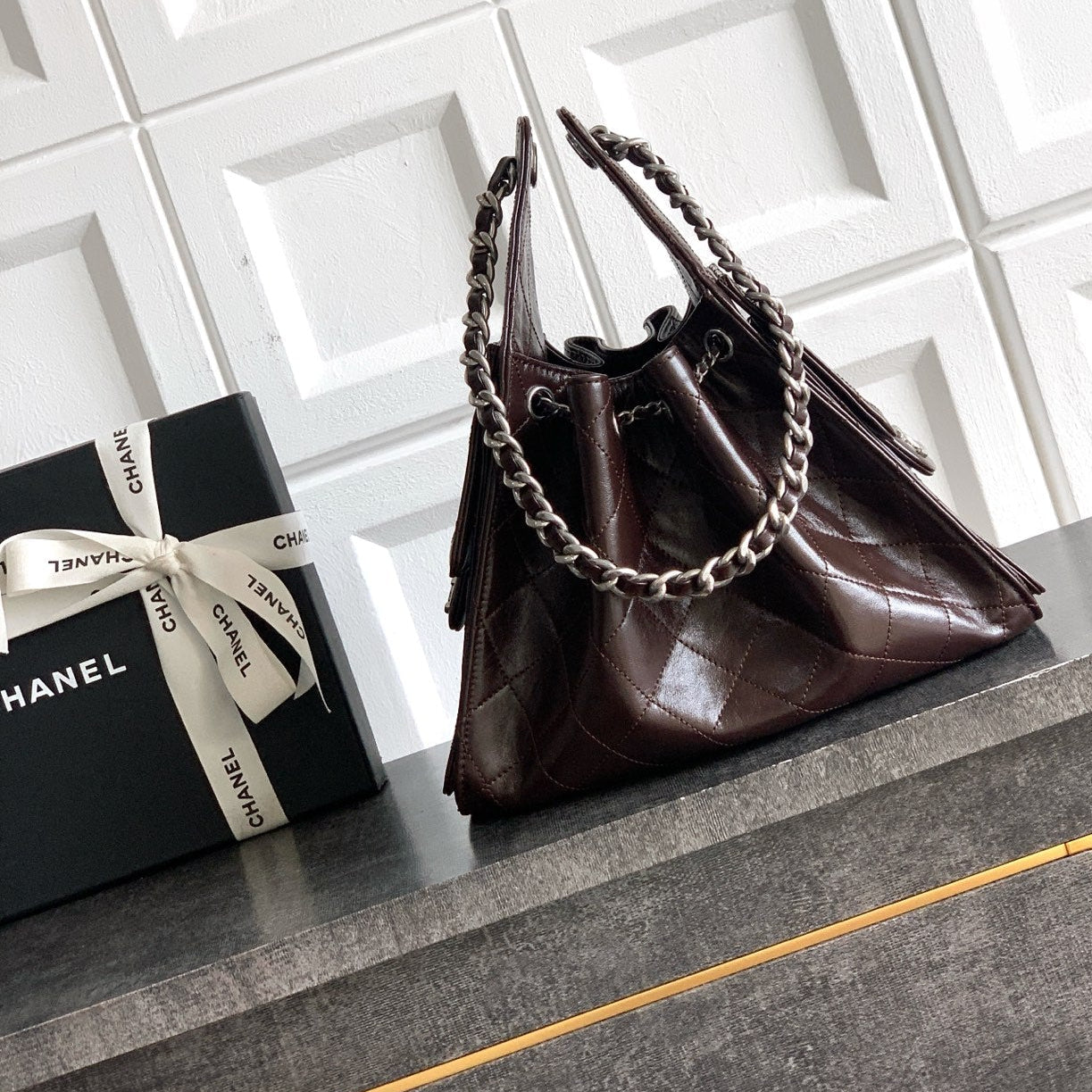 A dark brown Chanel handbag with silver chain straps, displayed on a gray cabinet next to a black Chanel gift box with a white ribbon bow against a white tiled wall.