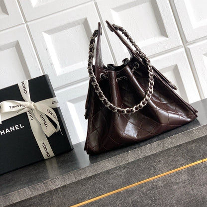 A dark brown Chanel handbag with silver chain straps, displayed on a gray cabinet next to a black Chanel gift box with a white ribbon bow against a white tiled wall.