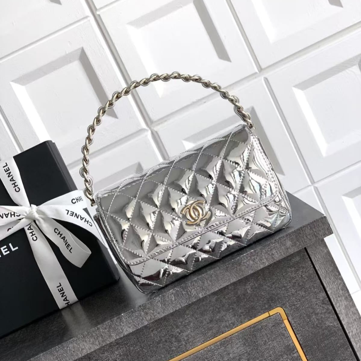 Silver quilted handbag with gold chain handle next to Chanel nail polish on a tiled surface