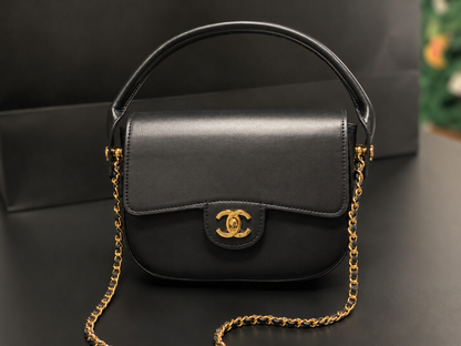 A black leather handbag with a gold-tone interlocking CC logo on the front flap, a top handle, and a gold chain strap, displayed on a dark surface.