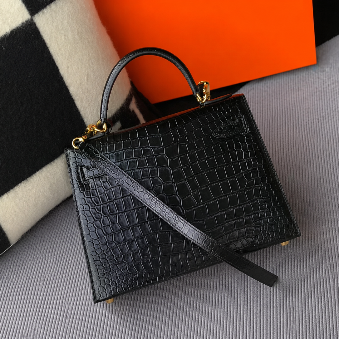 A structured black real crocodile-embossed leather handbag with a top handle and shoulder strap, styled on a gray fabric surface with a black-and-white patterned textile and an orange box in the background.