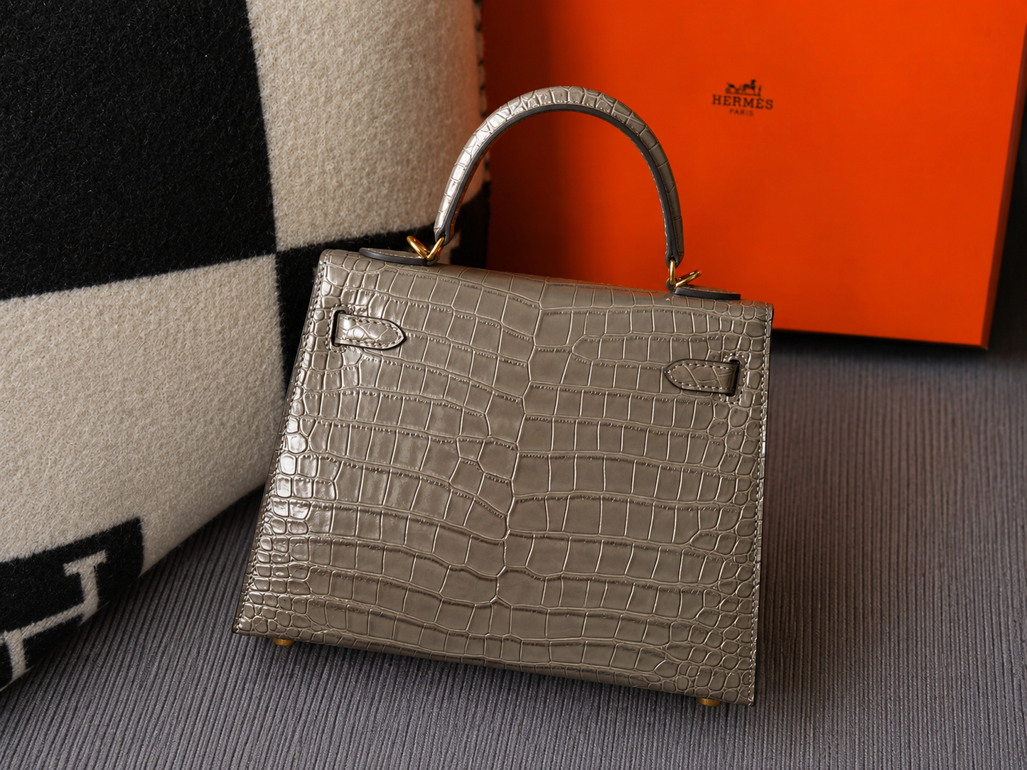 A structured mini handbag with a crocodile-embossed leather finish in taupe-gray, featuring gold-tone hardware and a top handle, styled with an orange Hermes box and a black-and-white checkered cushion in the background.